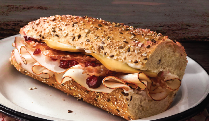 Publix sub on a white plate, bread with seeds and grains, filled with cheese, bacon and turkey.