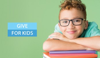 A boy leans on a small stack of colorful books smiling, with the words give for kids on the left.