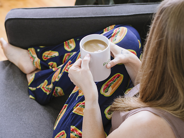 Young woman lounging on a grey couch drinking coffee in a white Publix mug wearing blue Publix sub pants