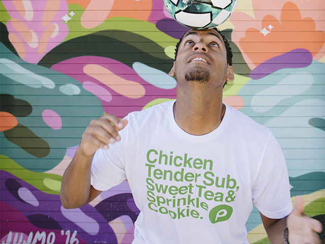 Young man balancing a colorful soccer ball on his head wearing a white Publix Chicken Tender Sub, Sweat Tea & Sprinkle Cookie t-shirt , in front of a colorful wall.